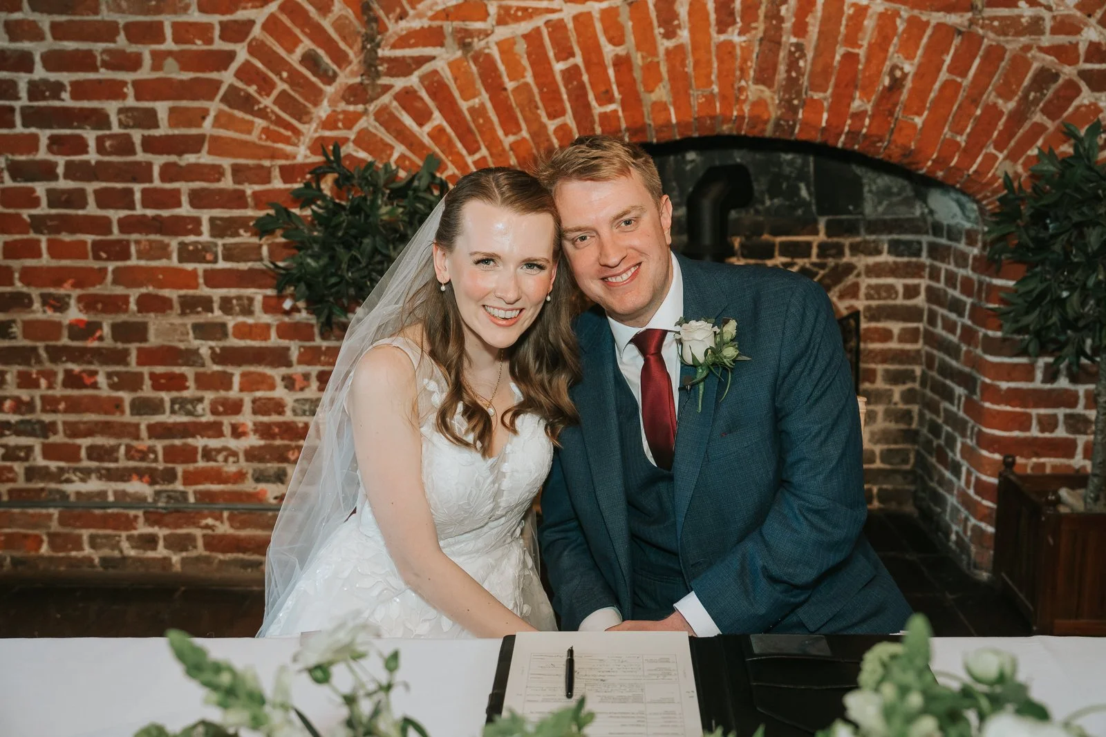  Newly married couple signing the register and smiling at the camera inside Tudor Barn Eltham with exposed brick fireplace and rustic Tudor barn wedding interiors. 