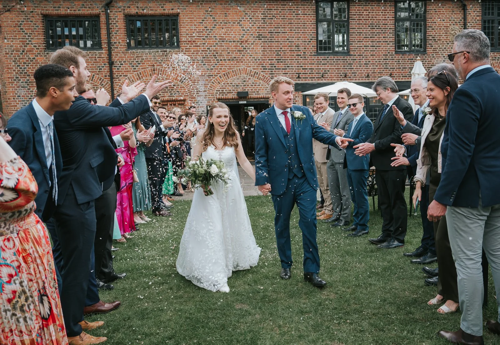  Bride and groom walking through a confetti tunnel outside Tudor Barn Eltham surrounded by cheering wedding guests in the historic Tudor barn courtyard. 