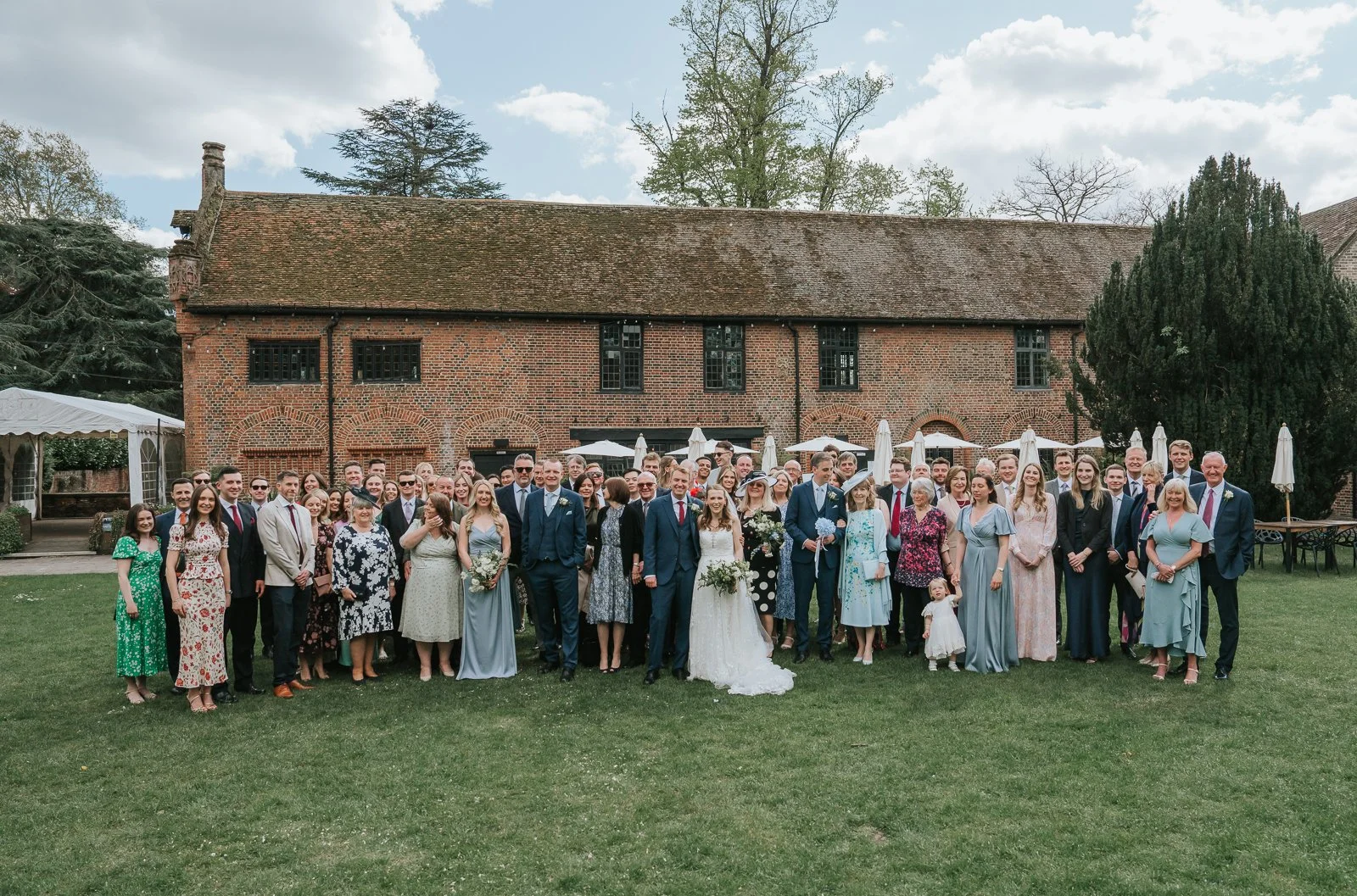  Large wedding group photo outside Tudor Barn Eltham with guests gathered on the lawn in front of the historic Tudor barn and beautiful brick facade. 