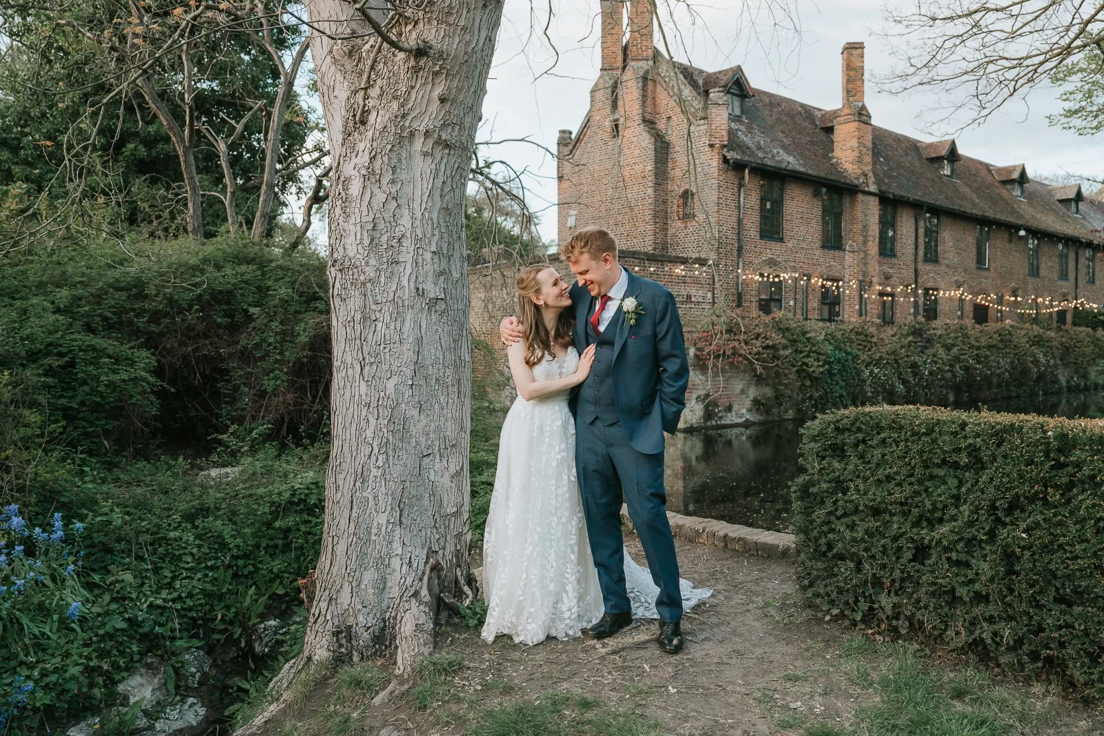  Bride and groom embracing beneath a tree in the romantic gardens at Tudor Barn Eltham during golden hour wedding portraits. 