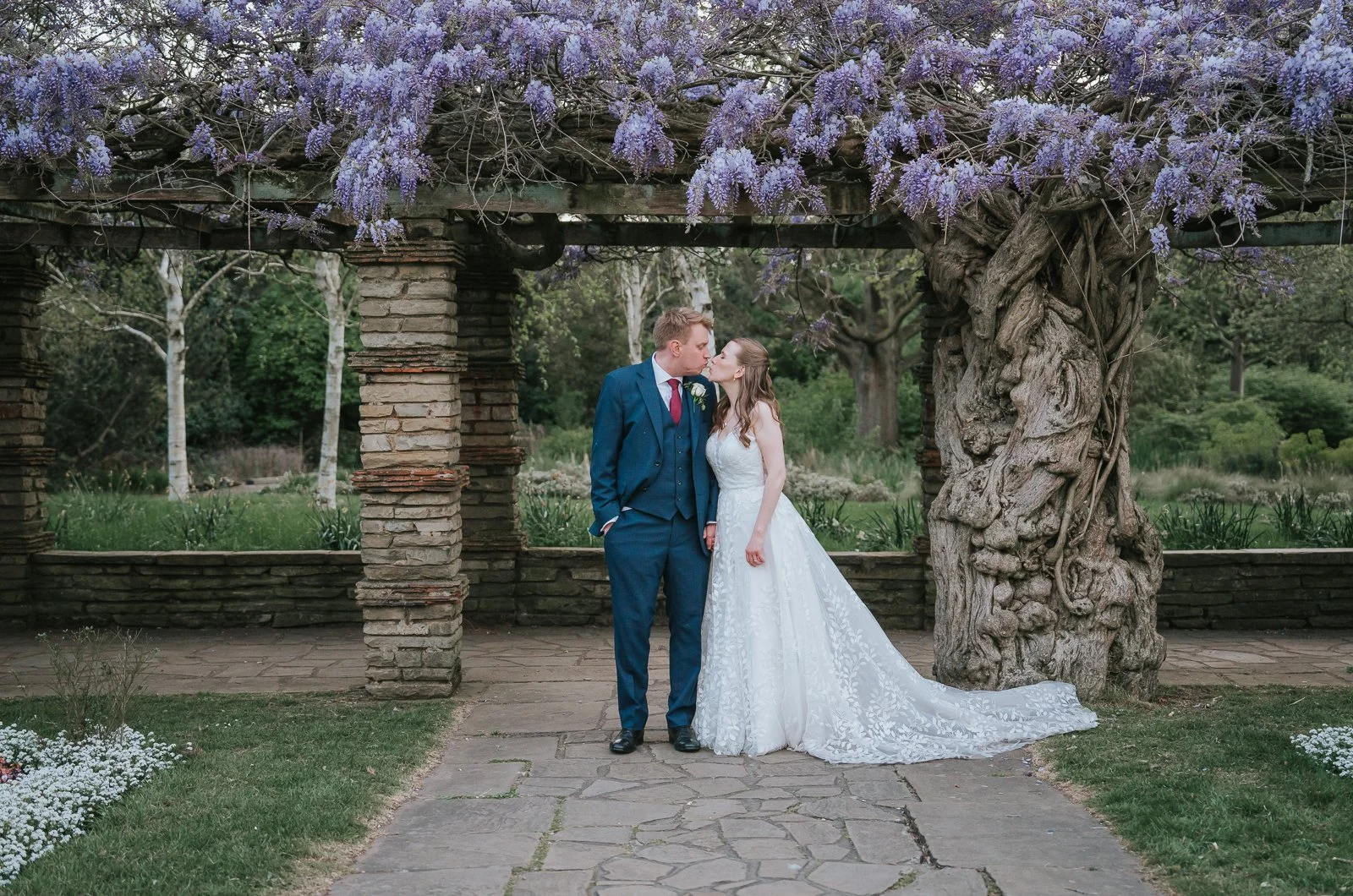  Newlyweds sharing a kiss beneath the wisteria-covered pergola in the gardens at Tudor Barn Eltham during their couples’ portraits. 