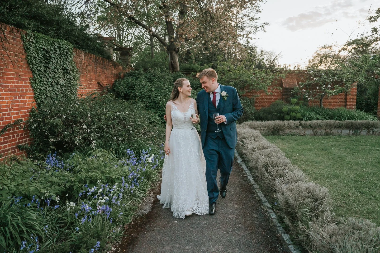  Bride and groom walking hand in hand through the formal gardens at Tudor Barn Eltham at sunset with champagne in hand. 