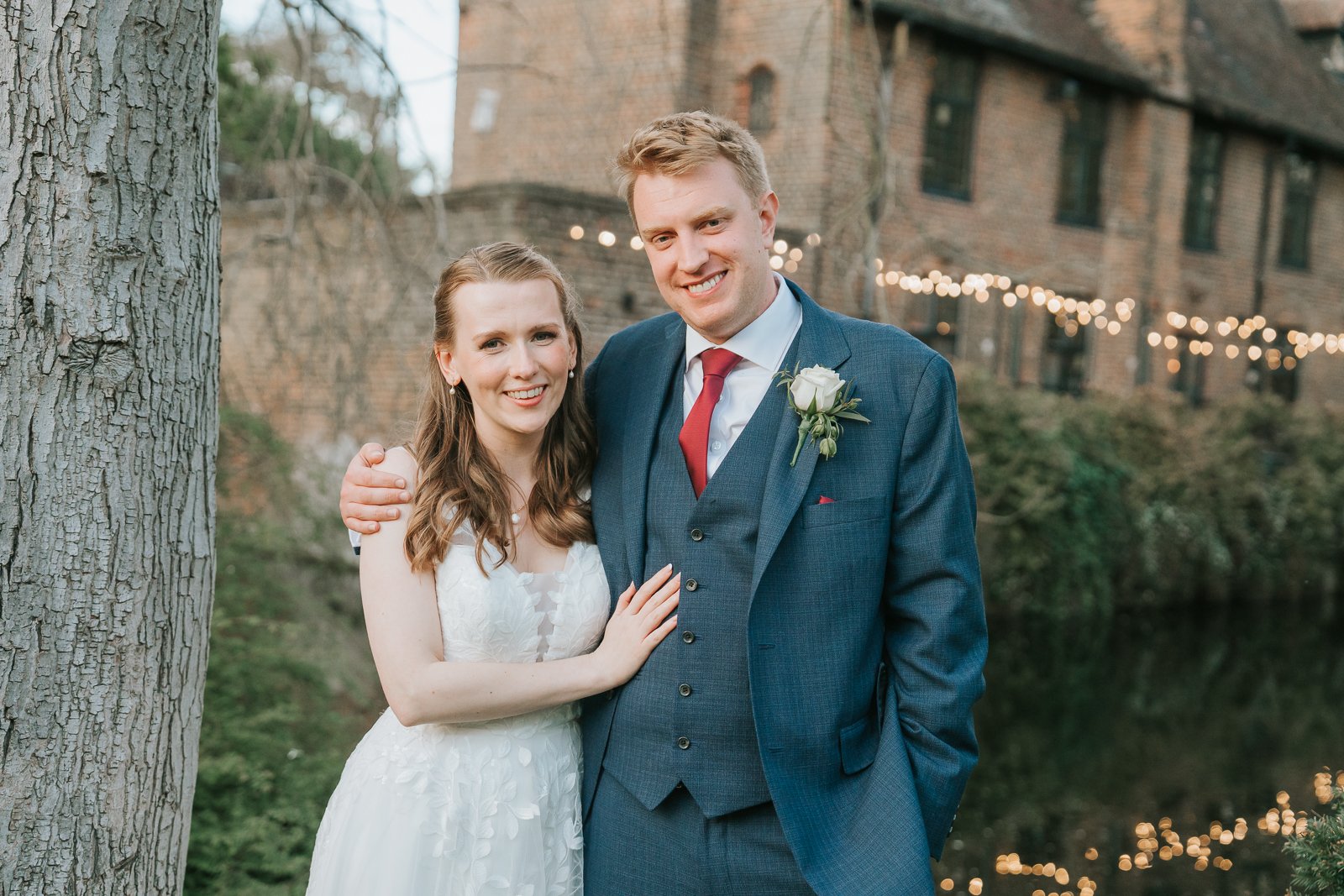  Natural wedding portrait of the newlyweds beside the moat at Tudor Barn Eltham with fairy lights and the historic barn in the background. 