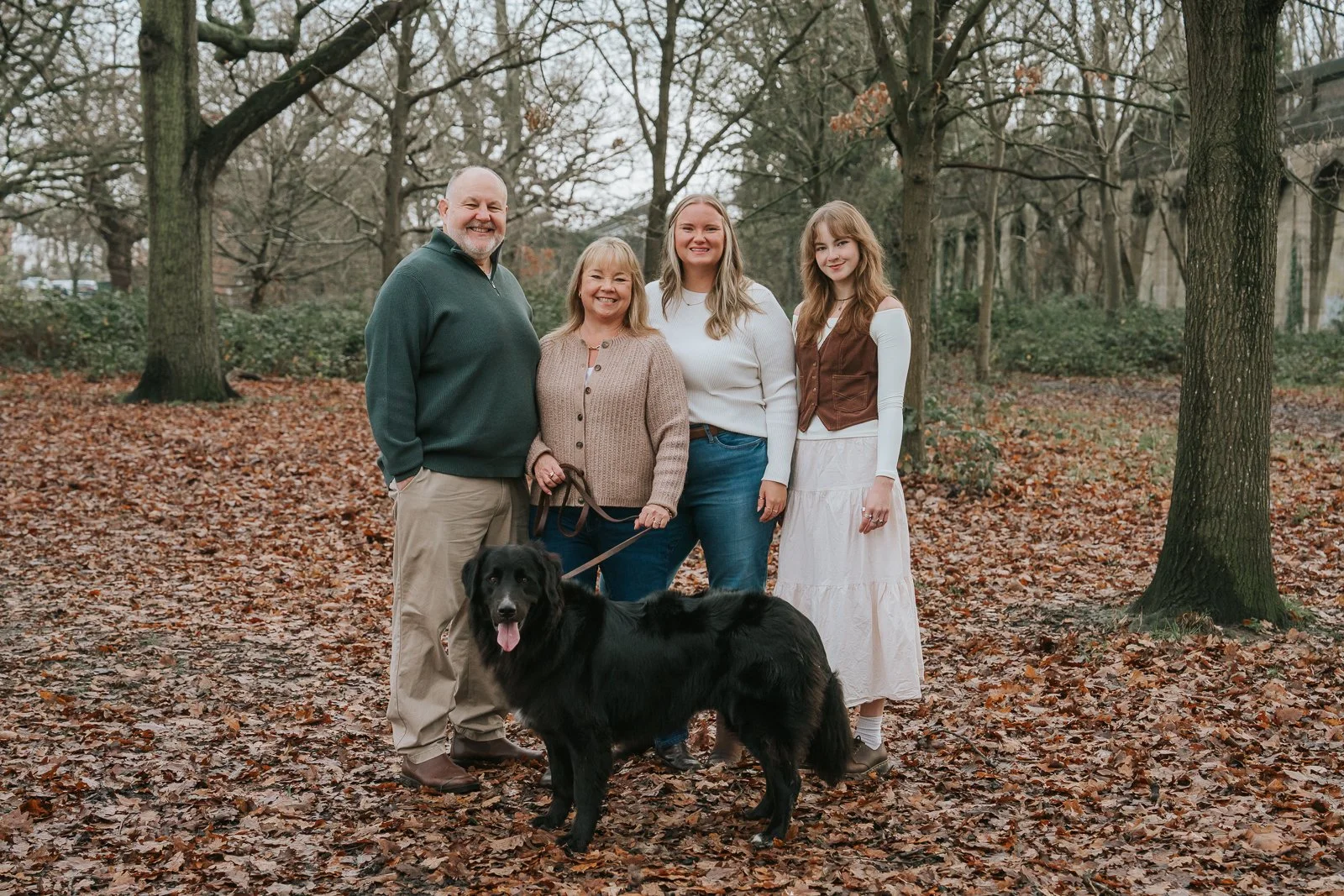  Family of four with their black dog during an autumn family photoshoot on Tooting Common. Natural outdoor family photography among fallen leaves and mature trees in South London. 