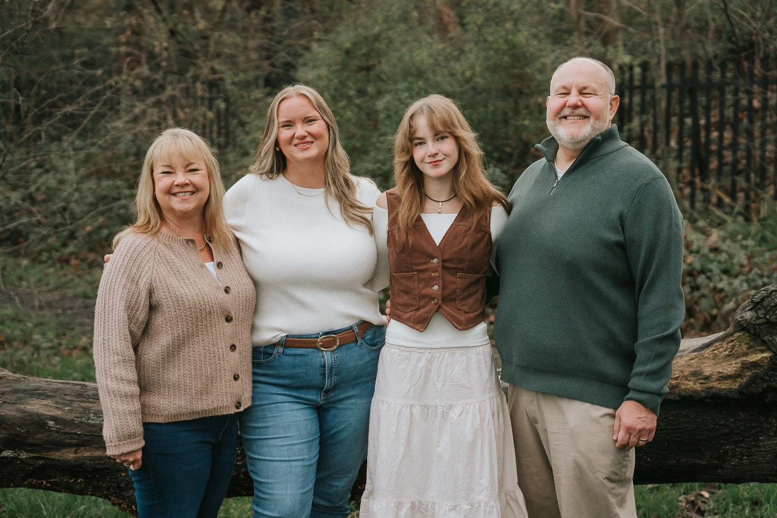  Family portrait on Tooting Common featuring parents and two grown-up daughters standing together beside a fallen tree, photographed in a natural woodland setting. Relaxed and natural family photography by a London family photographer. 
