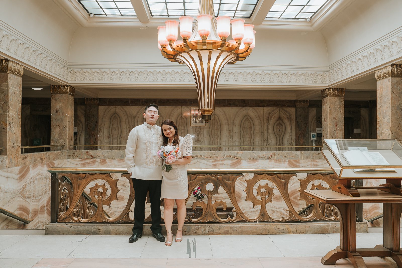  Newly married couple posing beneath the grand Art Deco chandelier in Wandsworth Town Hall's iconic marble atrium after their London registry office wedding. 