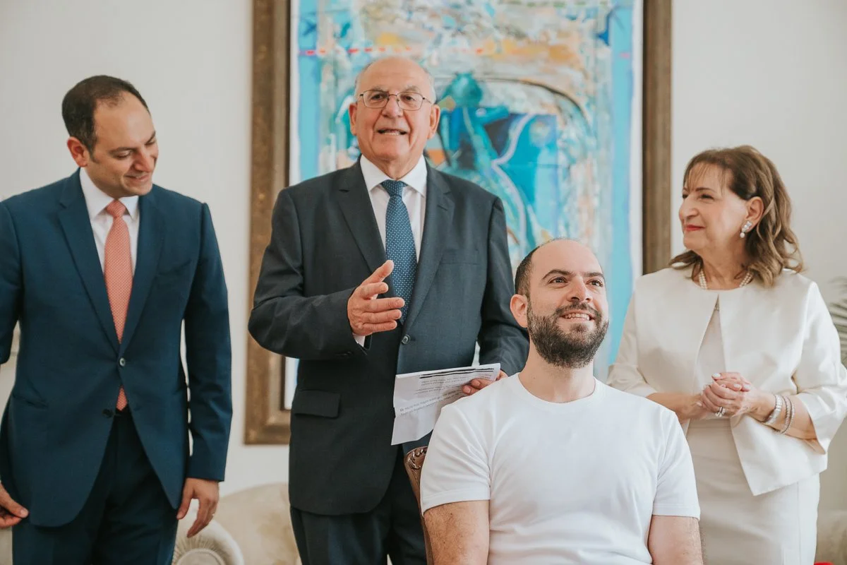  Greek Orthodox stolisma with the groom seated alongside family members as relatives gather to offer blessings and celebrate before the wedding ceremony. 
