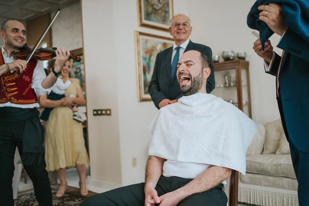  Joyful moment during a Greek Orthodox stolisma as the groom laughs while relatives and musicians participate in the traditional pre-wedding shaving ritual. 