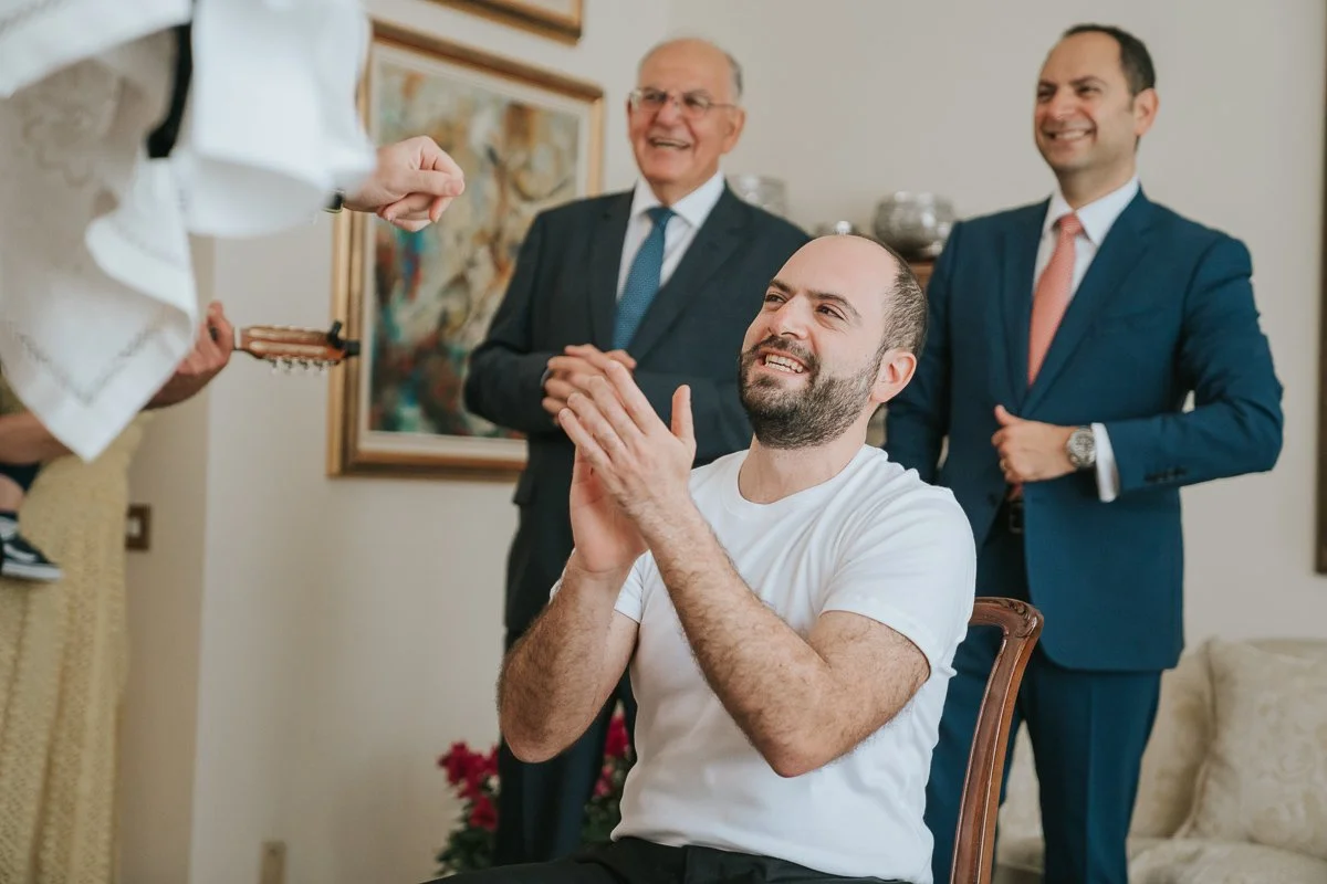  Groom applauding during his Greek wedding stolisma surrounded by family members, capturing the warmth and tradition of Orthodox wedding preparations in London. 