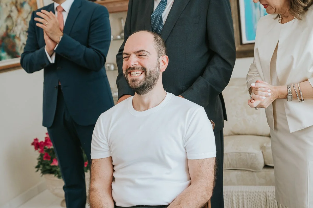  Groom smiling during his Greek wedding preparations surrounded by family members sharing in the joy and traditions of the stolisma. 