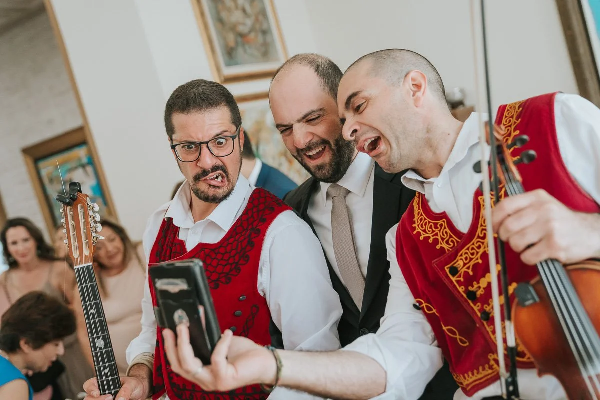  Groom laughing with traditional Greek musicians during a lively Orthodox stolisma, capturing the fun, music and family atmosphere of the pre-wedding celebrations before a Greek wedding in London. 