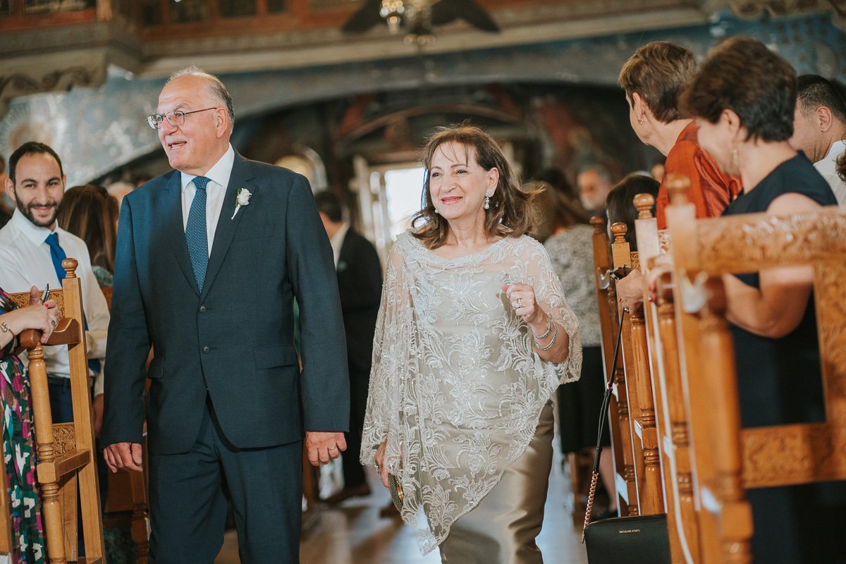  Parents of the groom arriving at a Greek Orthodox wedding ceremony, walking down the aisle of a historic Greek church surrounded by family and guests. 