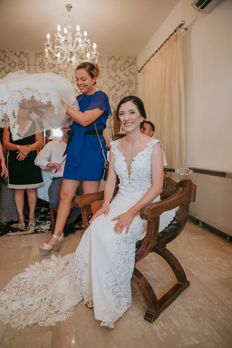  Bride smiling during her Greek Orthodox stolisma as family members prepare to place her veil during the traditional wedding preparations. 