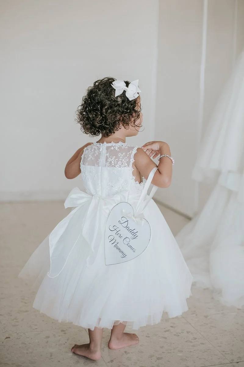  Flower girl in a white dress carrying a heart-shaped sign reading "Daddy Here Comes Mummy" during Greek wedding preparations. 