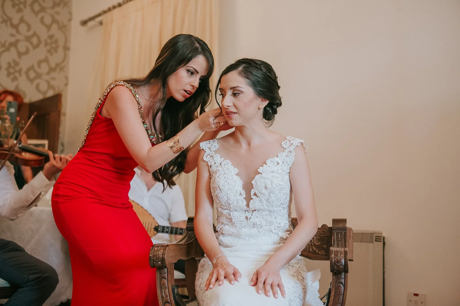  Bridesmaid adjusting the bride's earrings during Greek wedding preparations while traditional musicians perform in the background. 