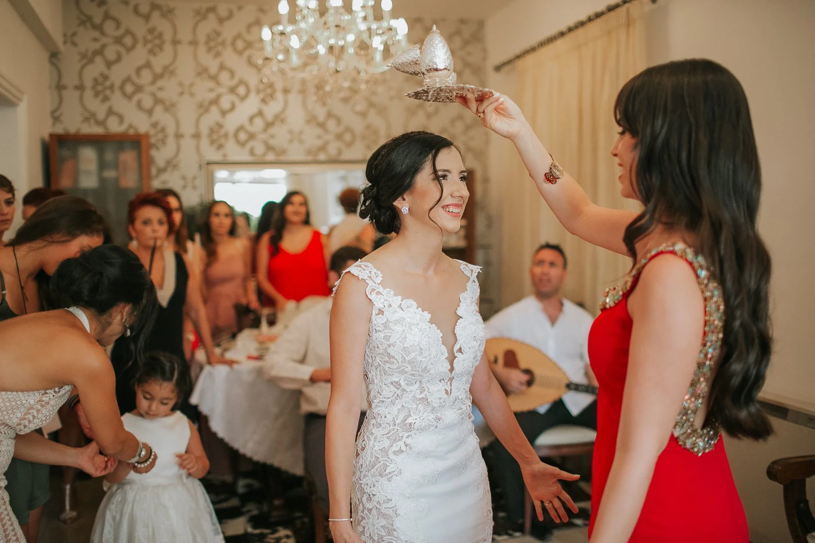  Bride smiling as a bridesmaid performs a traditional blessing during the stolisma, surrounded by family and friends celebrating her wedding day. 