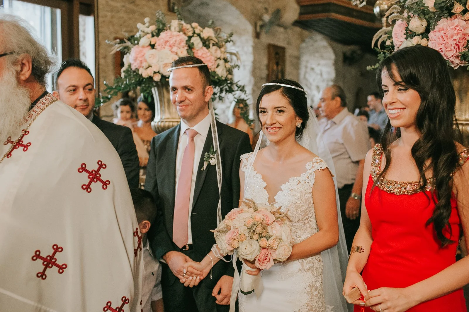  Bride and groom wearing stefana crowns during the sacrament of marriage in a Greek Orthodox church, surrounded by family and floral arrangements. 