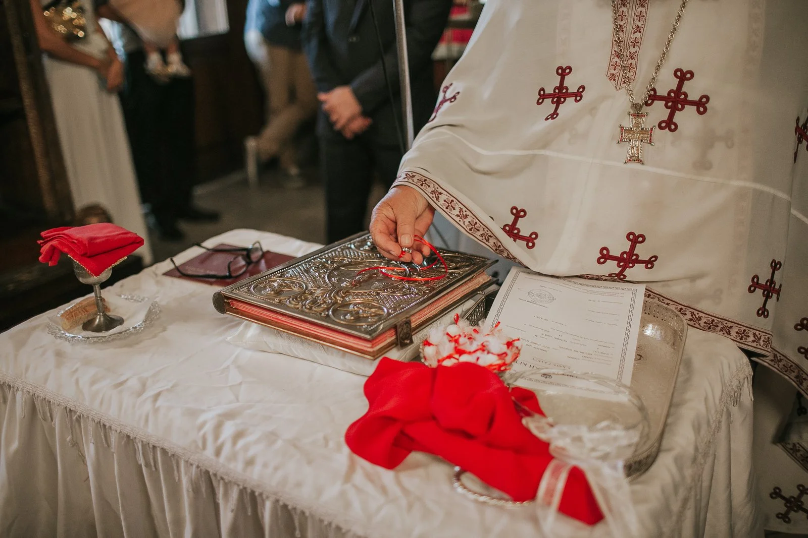  Greek Orthodox wedding rings resting on the Gospel book during the sacrament of marriage, with ceremonial red ribbons and silver church vessels on the altar table. 