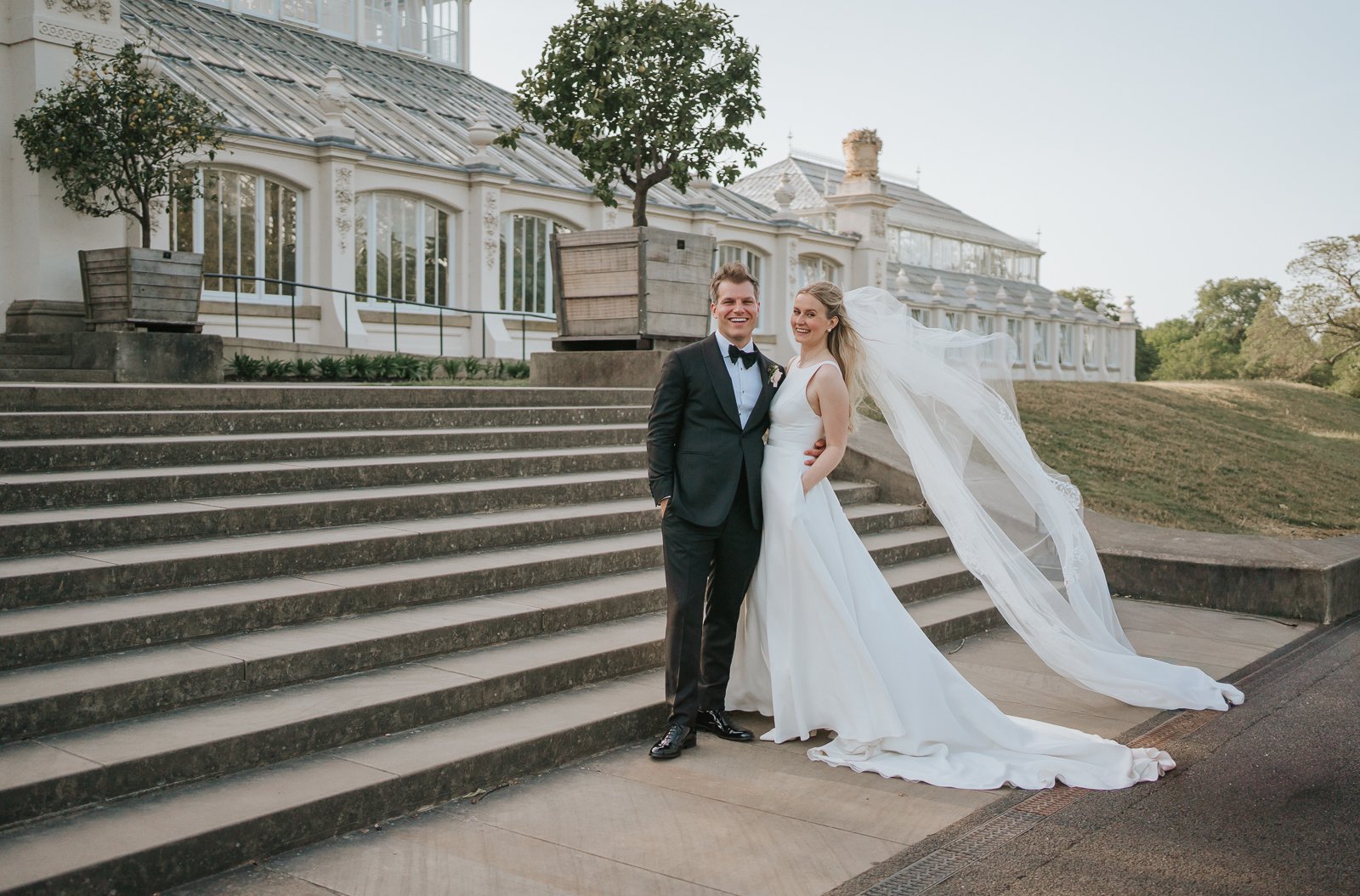  Bride and groom posing on the steps outside the Temperate House at Kew Gardens, with the bride’s veil flowing in the breeze during golden-hour wedding portraits. 
