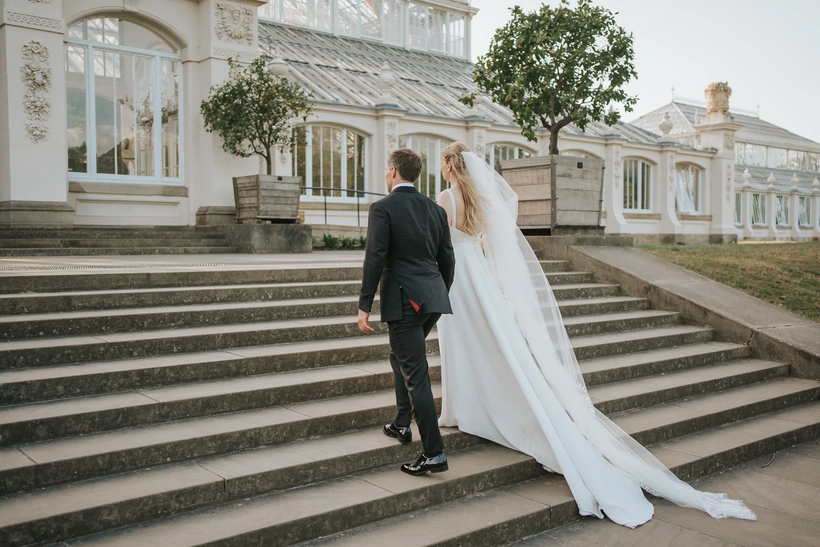  Bride and groom walking up the grand steps of the Temperate House at Kew Gardens after their wedding ceremony, with the elegant Victorian glasshouse behind them. 