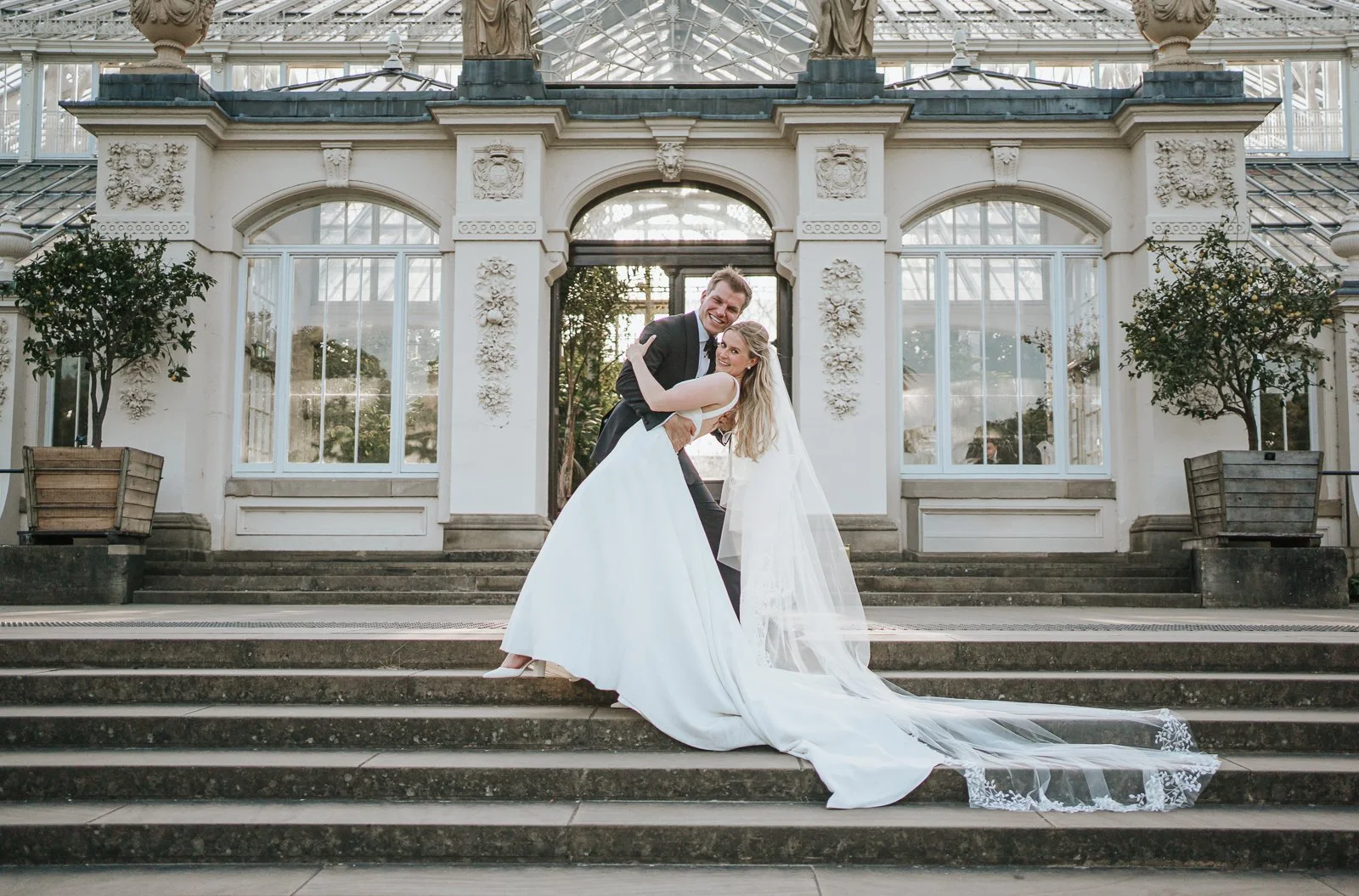  Fun and relaxed wedding portrait of a couple on the steps of the Temperate House at Kew Gardens, showcasing the venue’s stunning architecture and romantic setting for wedding photography. 