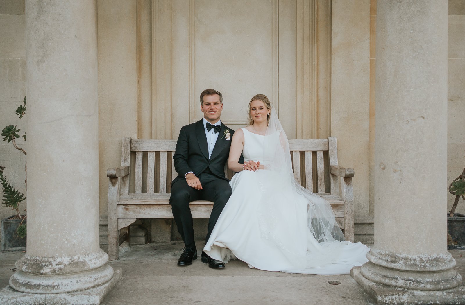  Bride and groom seated on a wooden bench beneath the classical columns of King William’s Temple at Kew Gardens, captured during relaxed wedding portraits. 