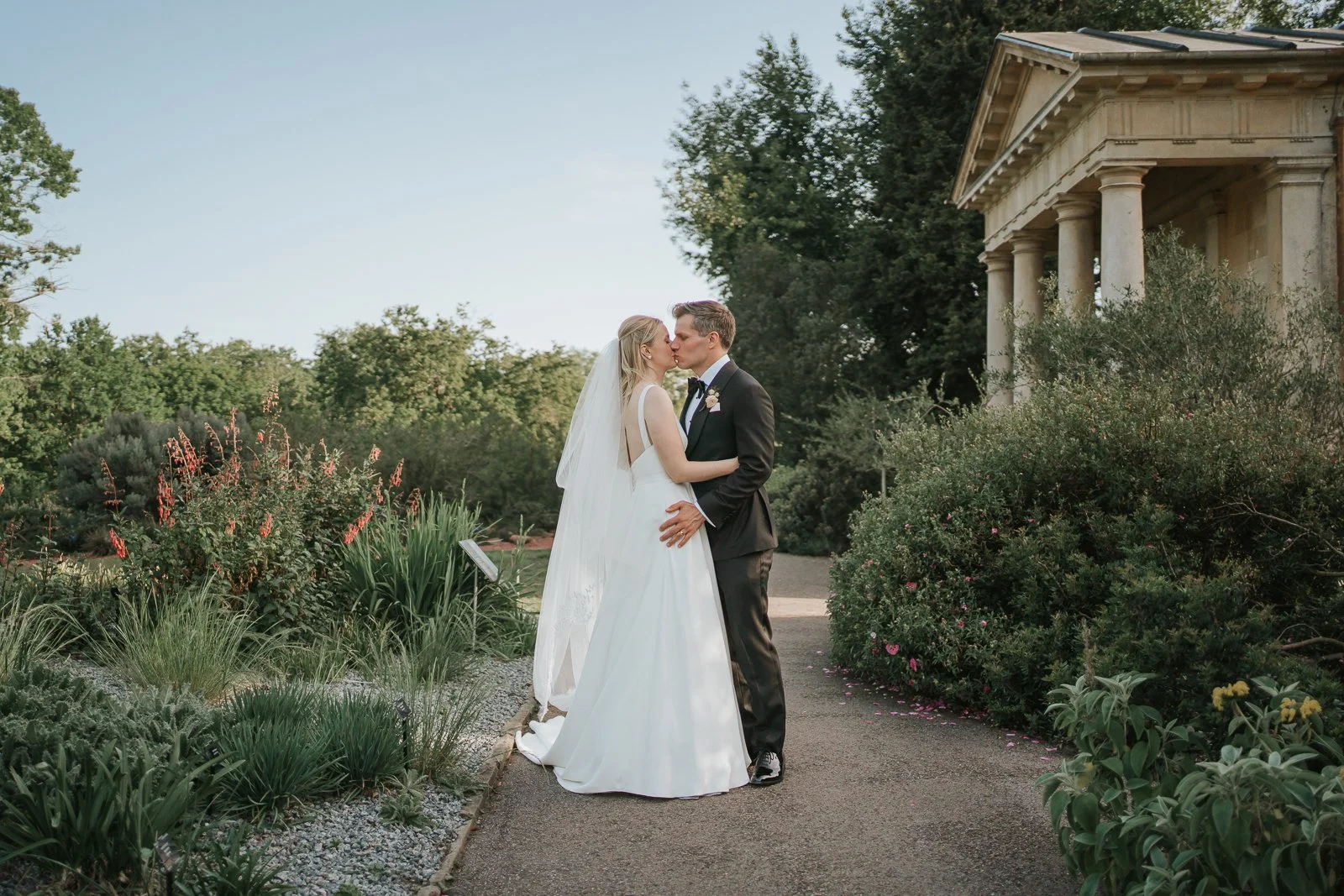  Bride and groom sharing a kiss along a garden pathway near King William’s Temple at Kew Gardens, surrounded by lush planting and seasonal flowers. 