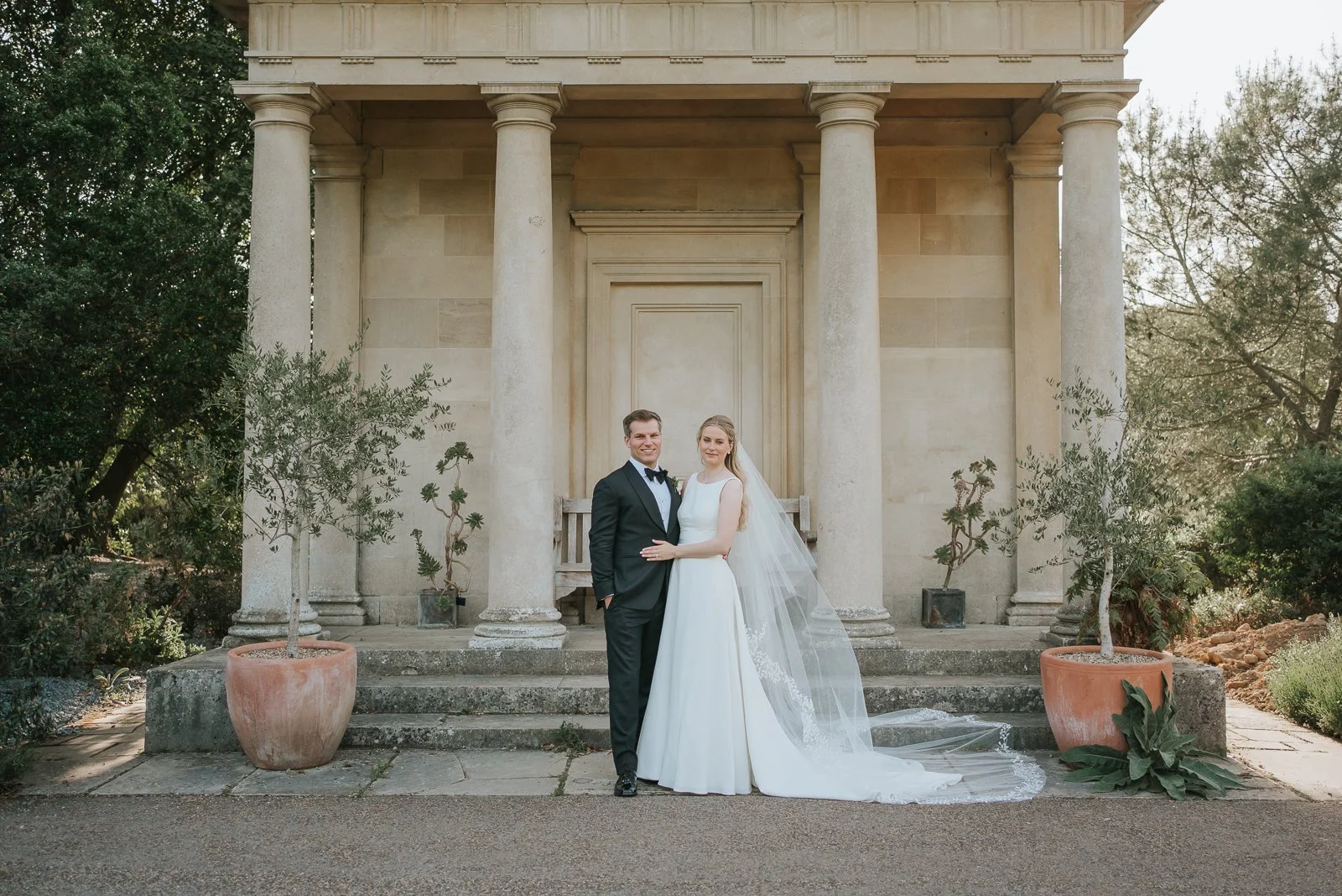  Bride and groom posing outside King William’s Temple at Kew Gardens, with the bride’s long veil flowing across the steps of the historic pavilion. 