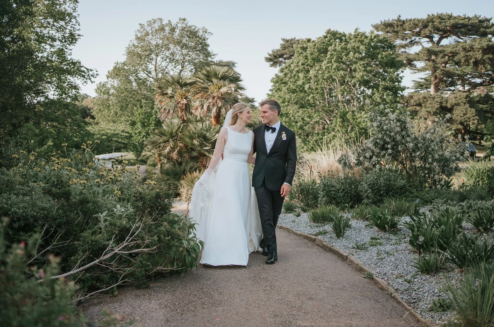  Bride and groom walking hand in hand through the Mediterranean Garden at Kew Gardens, surrounded by lush planting and exotic trees during relaxed wedding portraits. 