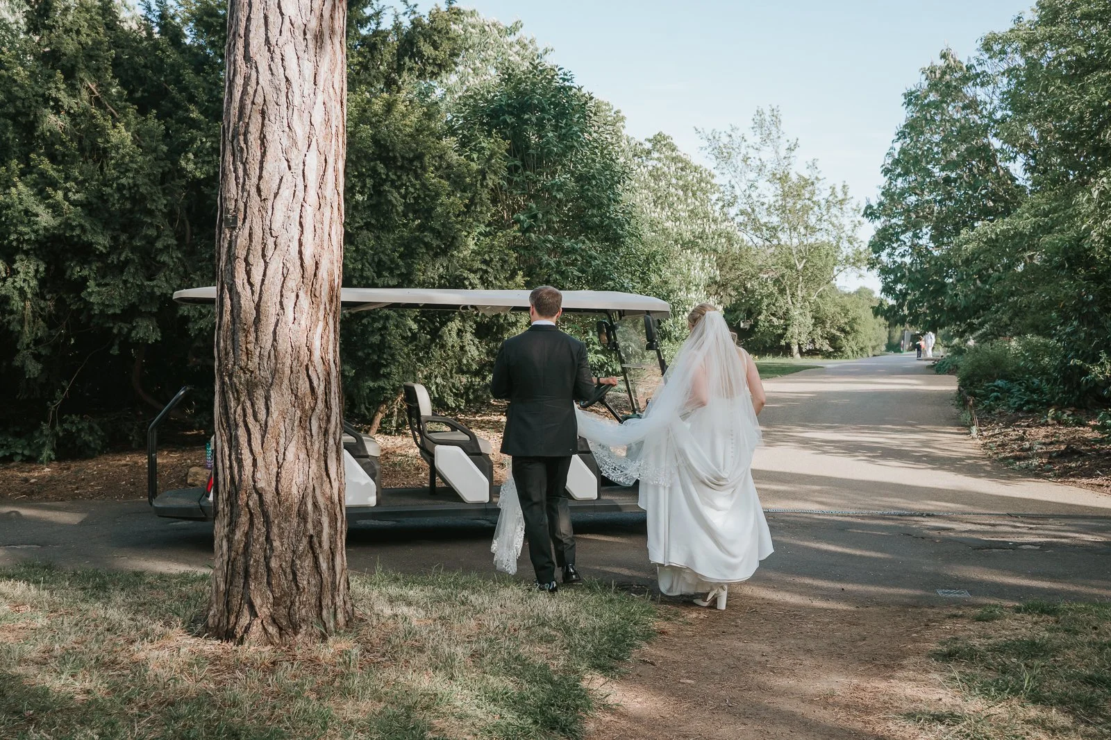  Bride and groom walking hand in hand through Kew Gardens after their wedding ceremony, making their way towards a golf buggy beneath the trees. 
