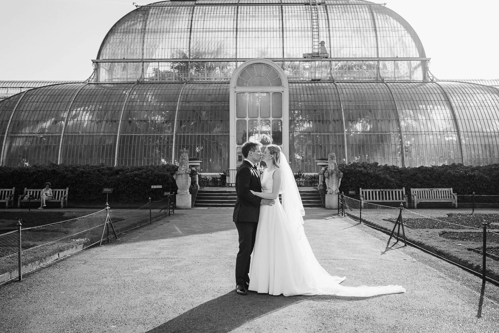  Black and white wedding portrait of the newlyweds embracing in front of The Palm House at Kew Gardens, highlighting the timeless architecture and romantic atmosphere. 