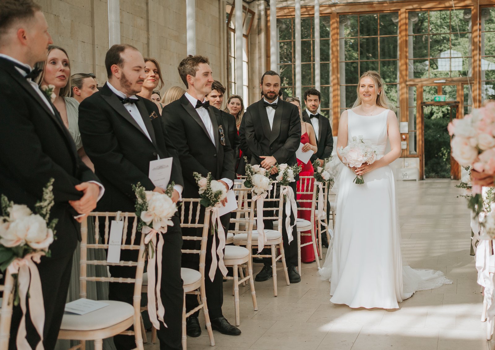  Bride walking down the aisle inside the Nash Conservatory at Kew Gardens, surrounded by floral arrangements and wedding guests. 