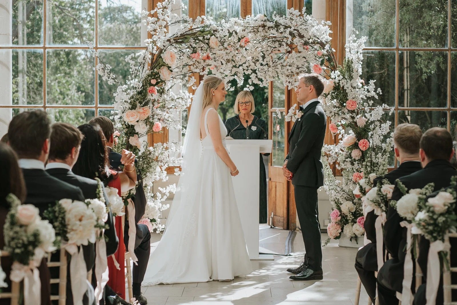  Wedding ceremony inside the Nash Conservatory at Kew Gardens, with the couple exchanging vows beneath a floral arch surrounded by family and friends. 