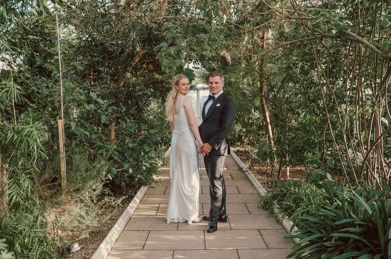  Bride and groom walking hand in hand through the lush tropical planting inside the Temperate House at Kew Gardens. 