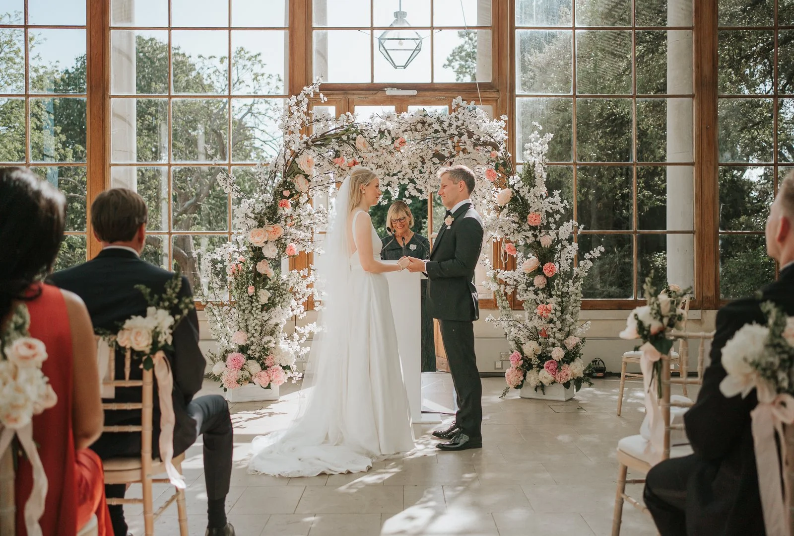  Bride and groom holding hands beneath a floral ceremony arch during their wedding ceremony in the Nash Conservatory at Kew Gardens. 