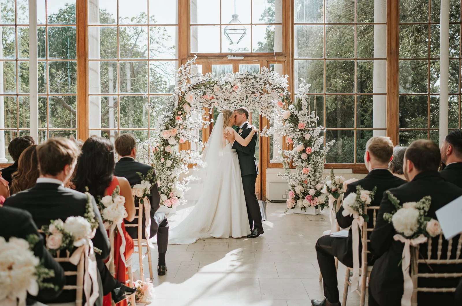  Bride and groom holding hands beneath a floral ceremony arch during their wedding ceremony in the Nash Conservatory at Kew Gardens. 