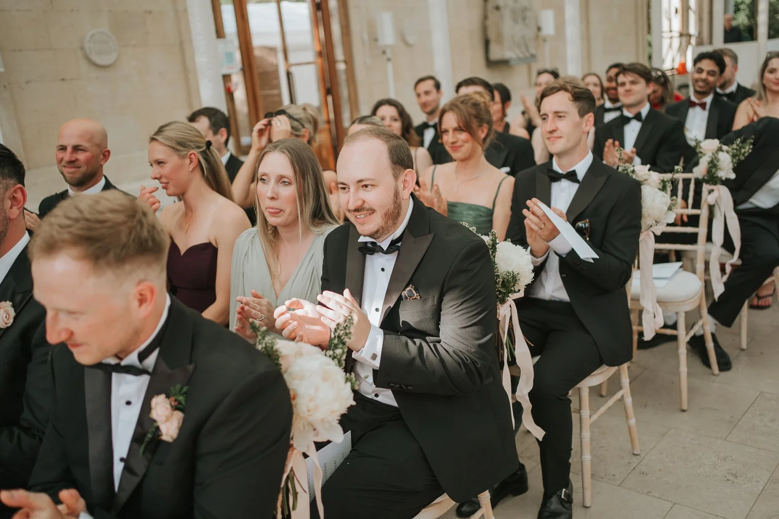  Wedding guests applauding during the ceremony inside the Nash Conservatory, surrounded by elegant floral aisle decorations. 