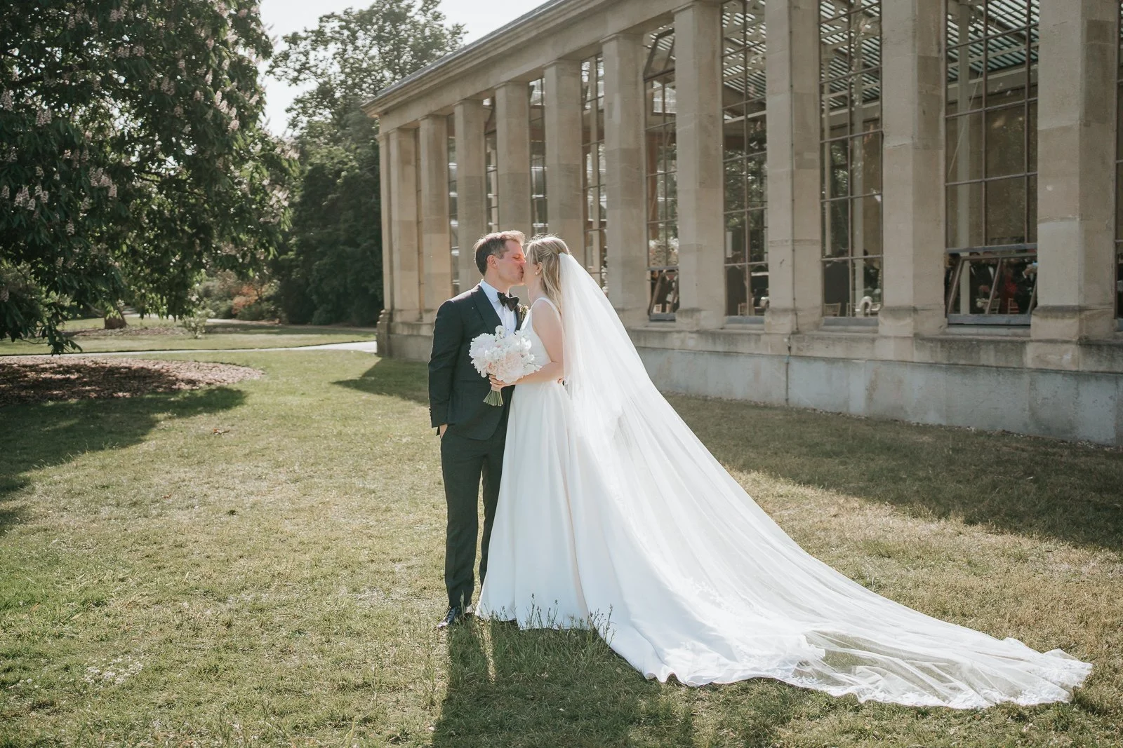  Bride and groom sharing a kiss outside the Nash Conservatory at Kew Gardens, with the bride’s long veil flowing across the lawn. 