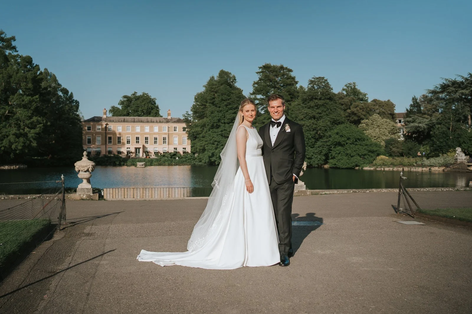 Bride and groom portrait beside the lake at Kew Gardens, with the historic mansion and landscaped grounds in the background. 