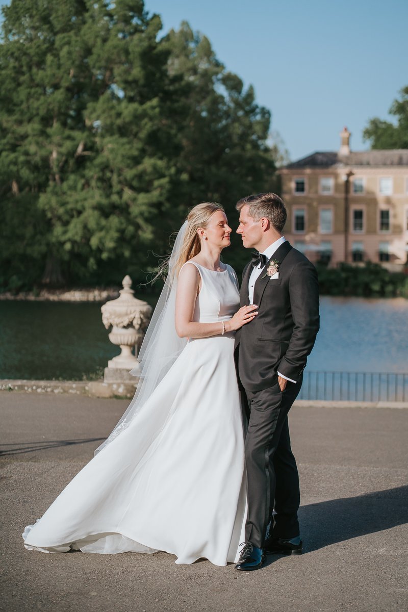  Bride and groom sharing a quiet moment beside the lake at Kew Gardens, with the historic mansion and gardens creating a romantic backdrop for their kew gardens wedding photography. 
