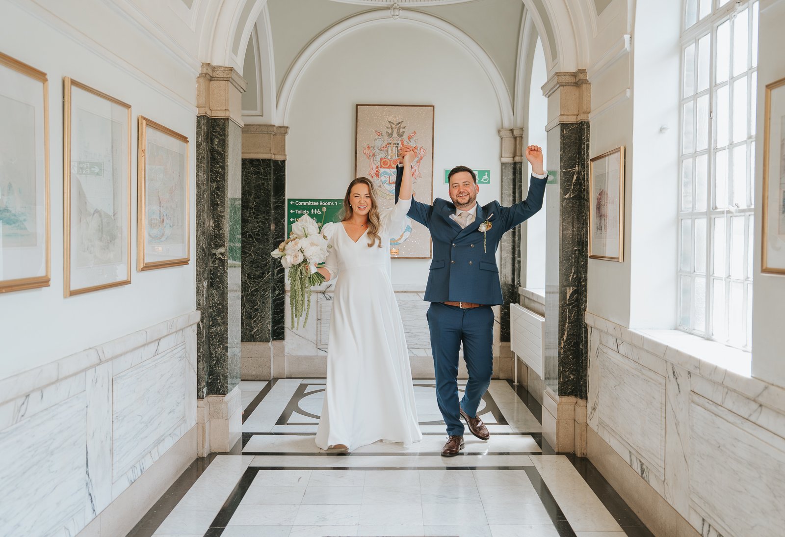  Newlyweds celebrating with raised hands in the grand marble hallway of Islington Town Hall. 