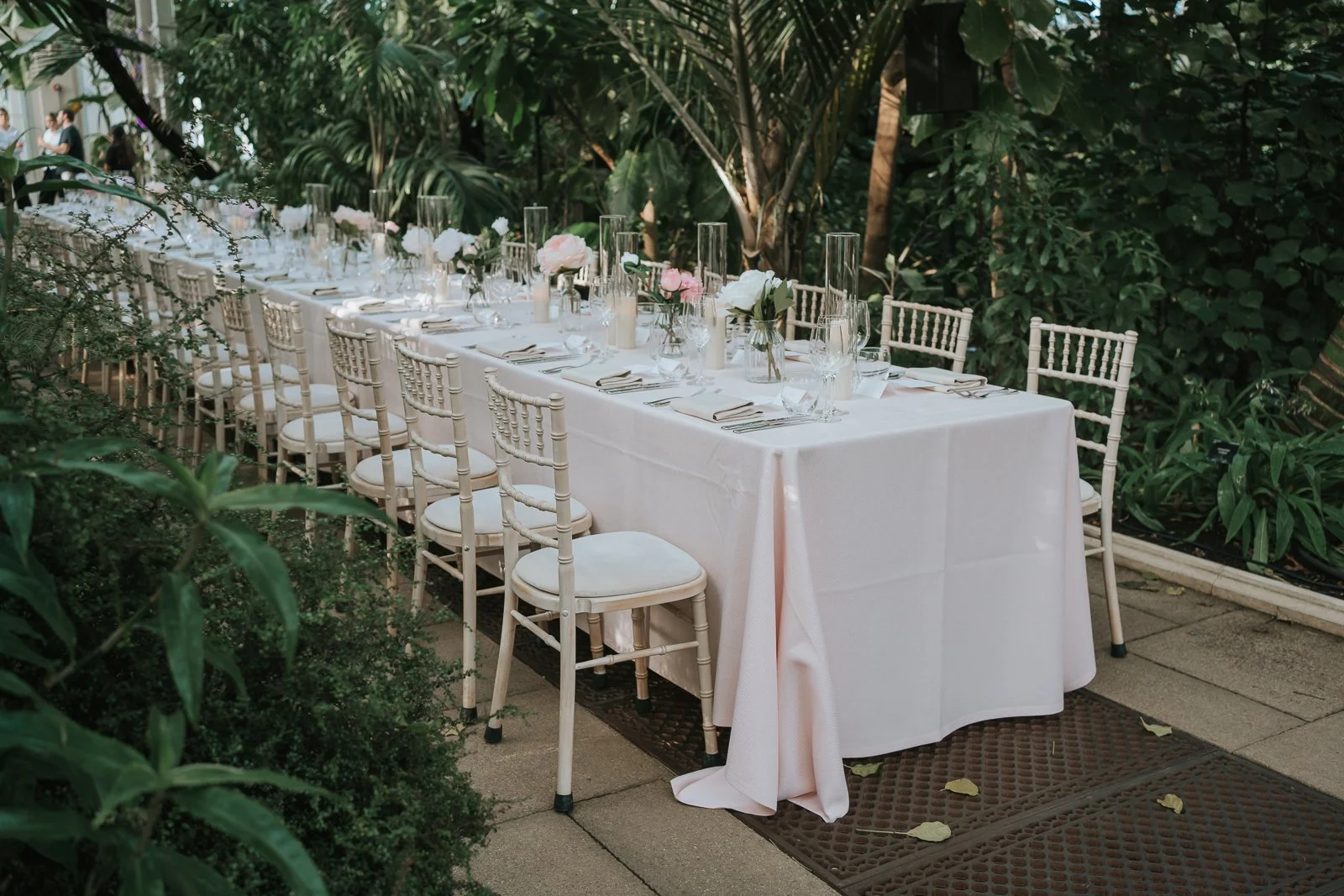  Elegant wedding breakfast table with pale pink linens, floral centrepieces and Chiavari chairs nestled amongst the greenery of the Temperate House at Kew Gardens. 