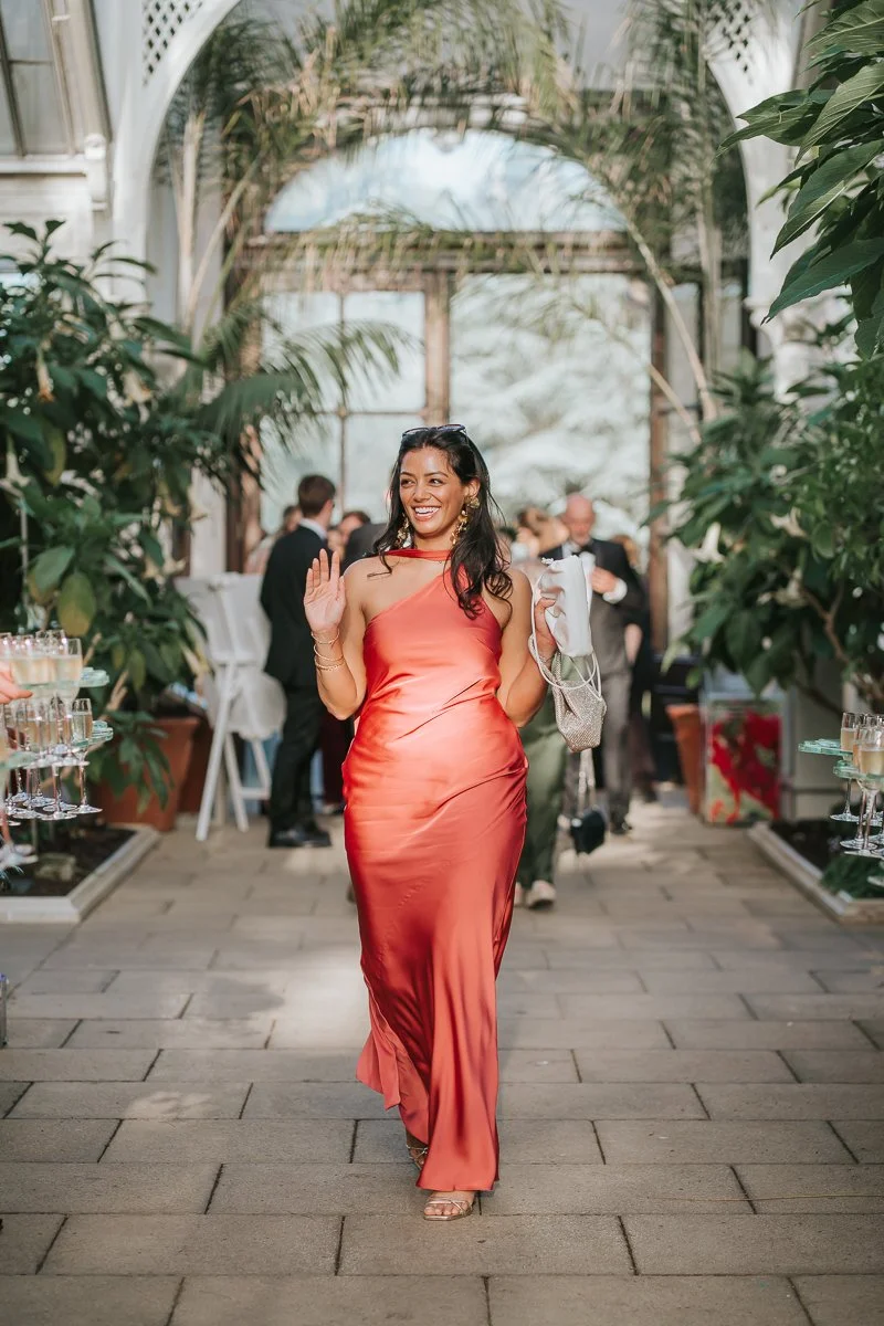  Wedding guest arriving at the Temperate House reception at Kew Gardens, surrounded by tropical plants and beautiful Victorian glasshouse architecture. 
