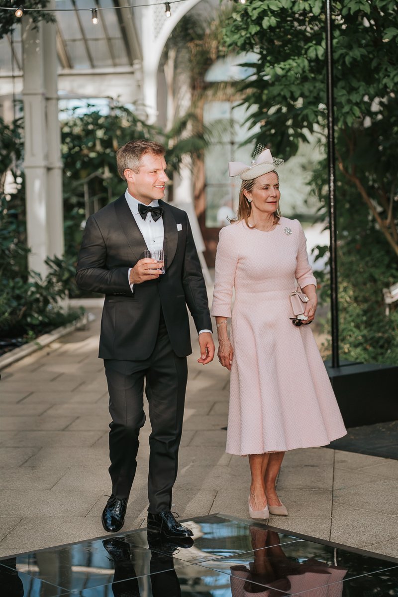  Wedding guests strolling through the Temperate House at Kew Gardens during an evening reception amongst exotic plants and soft natural light. 