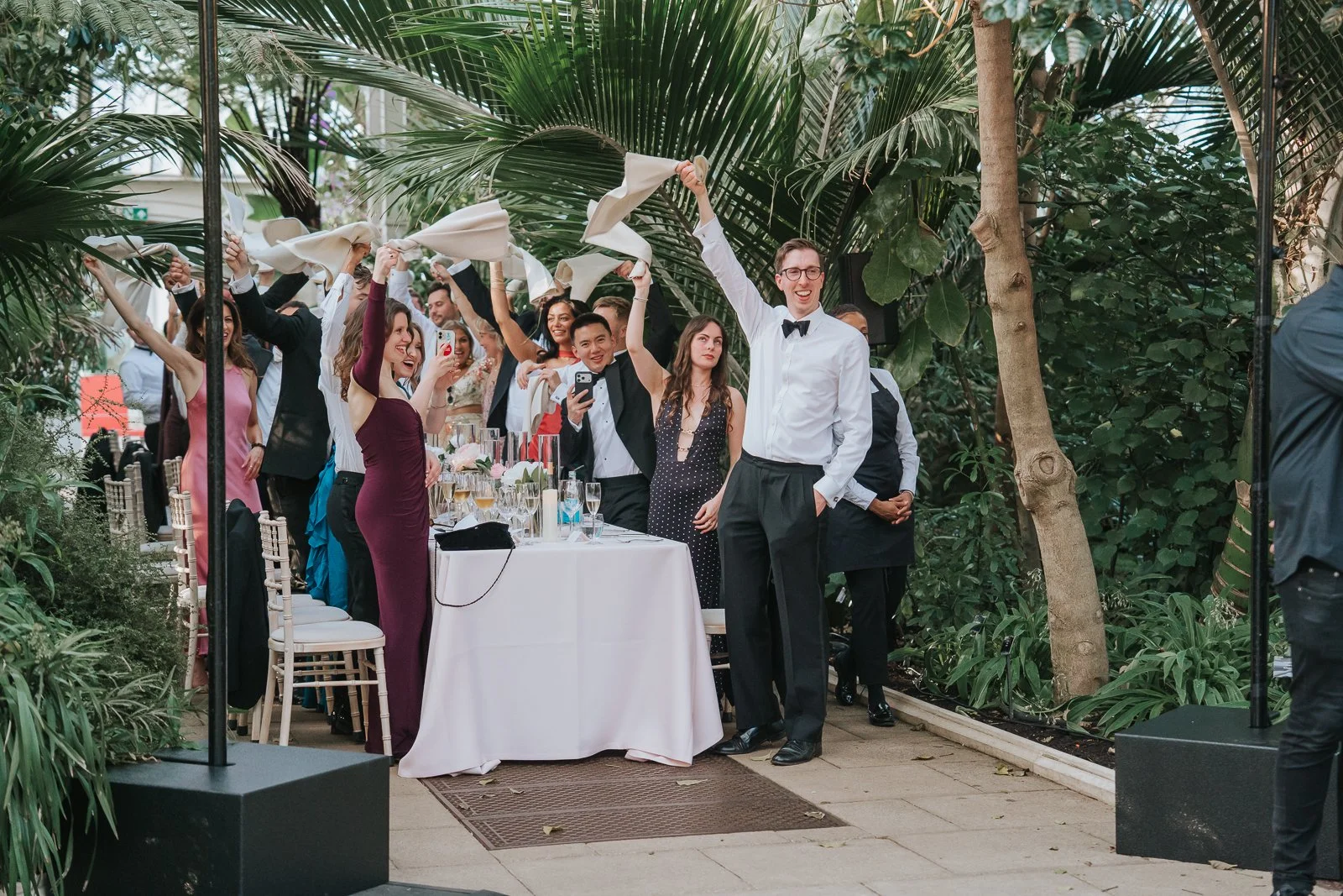  Wedding guests waving napkins and celebrating during the wedding breakfast in the Temperate House at Kew Gardens, creating a fun and lively atmosphere. 