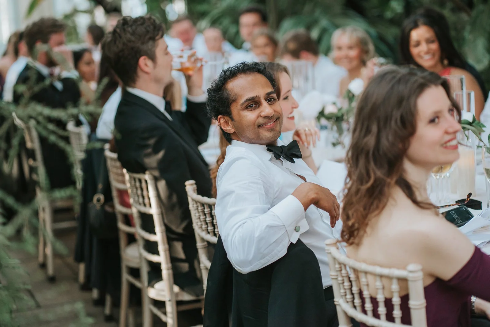  Wedding guests enjoying speeches and laughter during a reception in the Temperate House at Kew Gardens, seated amongst exotic plants and candlelit tables. 