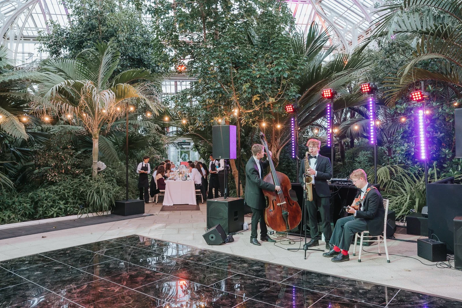  Wedding guest delivering a speech on the dance floor during a Temperate House wedding reception at Kew Gardens, surrounded by tropical plants, elegant catering displays and the iconic Victorian glasshouse. 