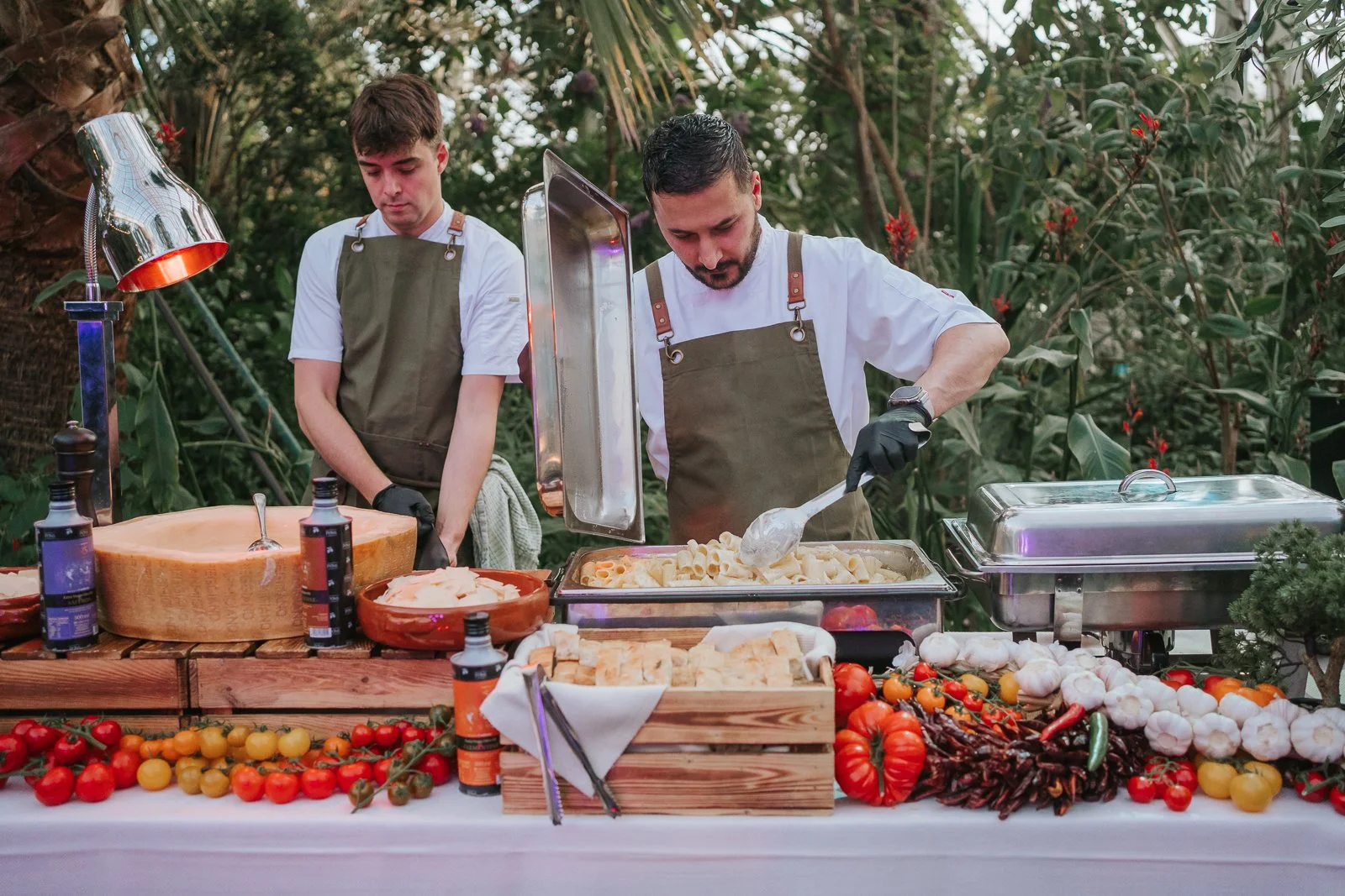  Wedding caterers serving fresh pasta from a live food station during a reception in the Temperate House at Kew Gardens, surrounded by colourful seasonal produce. 