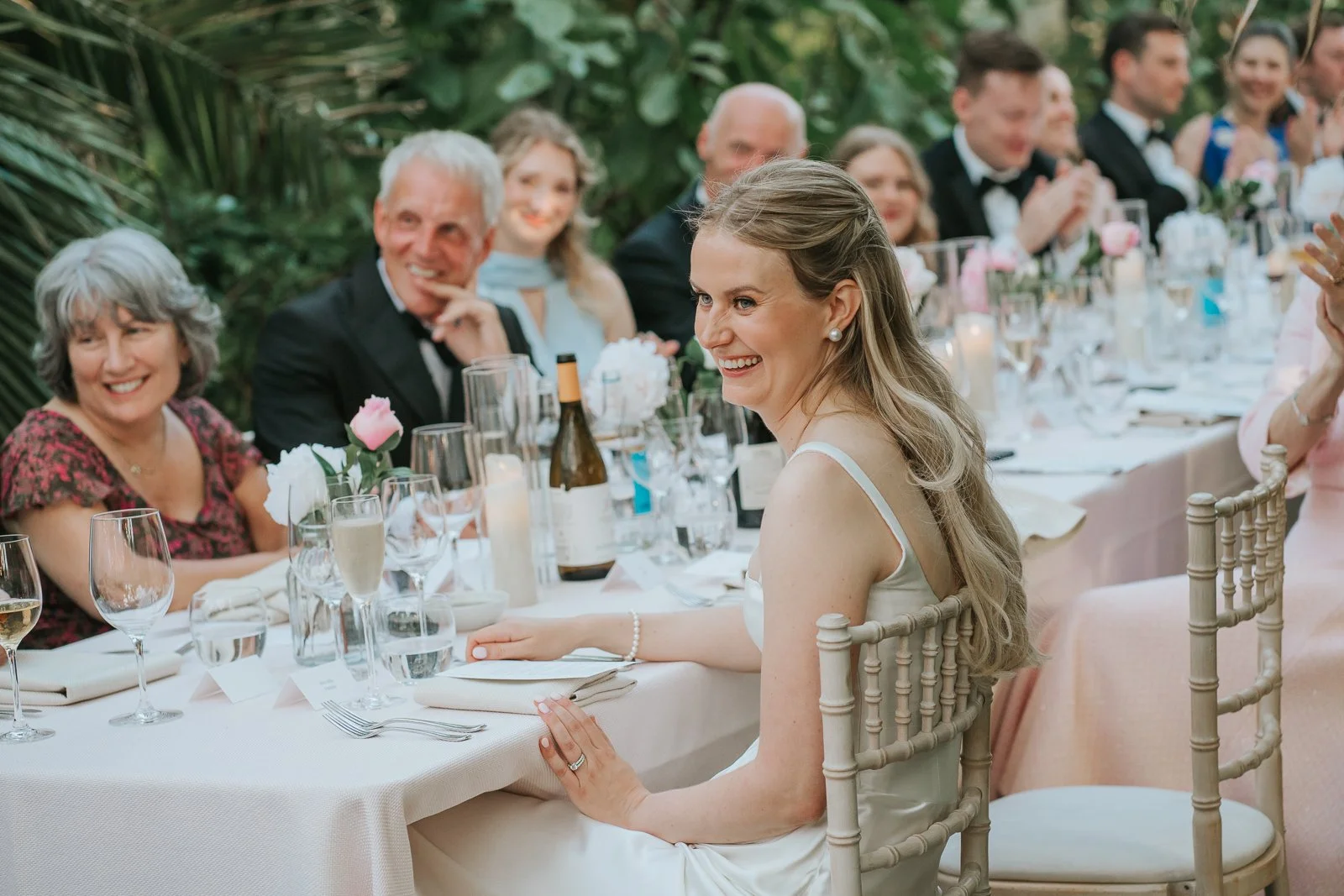  Bride smiling during wedding speeches inside the Temperate House at Kew Gardens, with family and friends gathered around the reception table. 