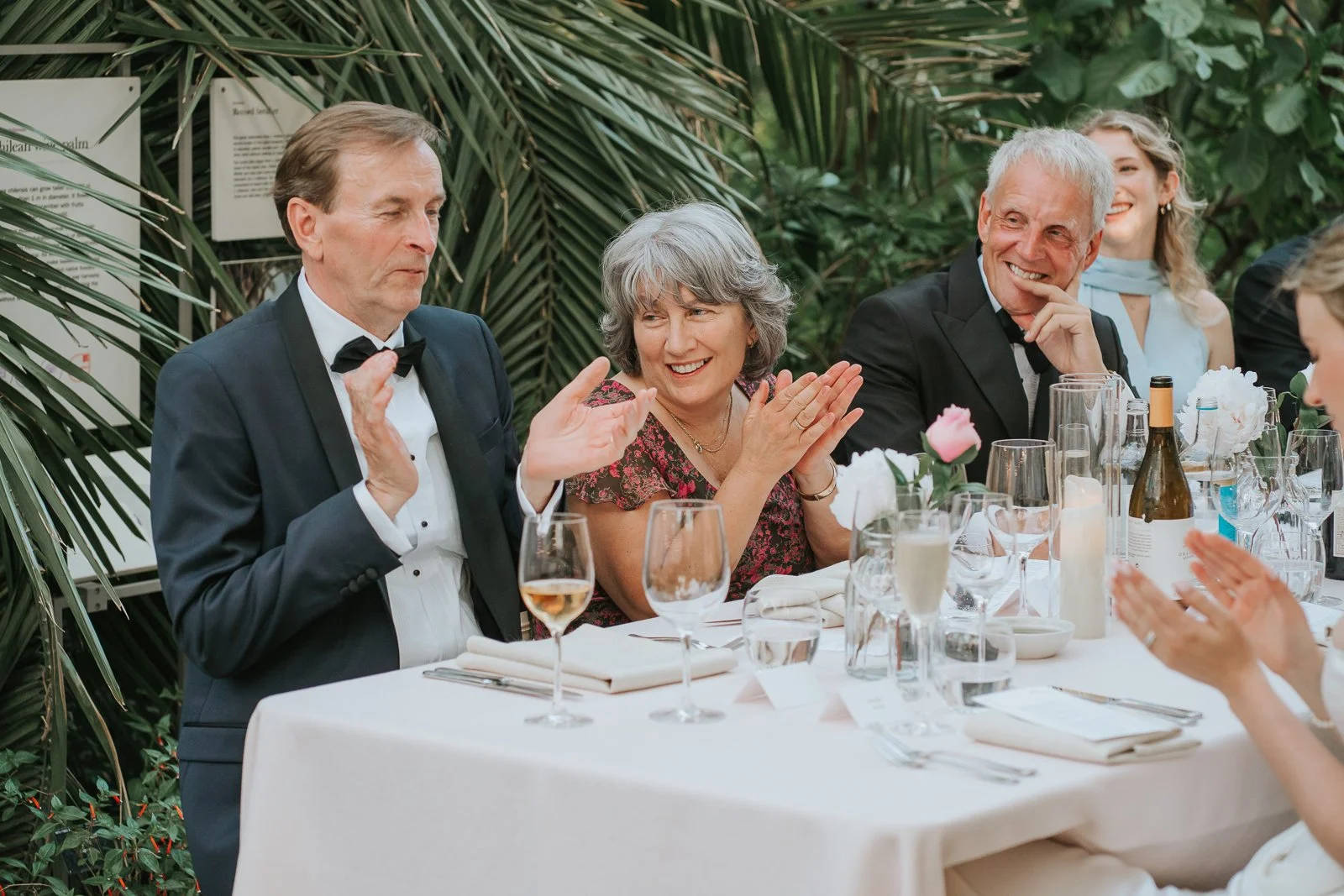  Wedding guests applauding and smiling during wedding speeches in the Temperate House at Kew Gardens, gathered around elegantly styled reception tables surrounded by lush tropical plants. 